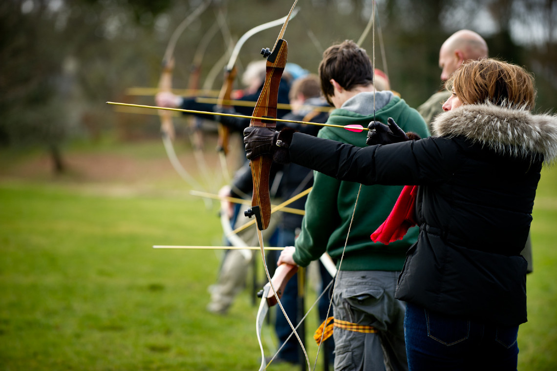 a group of adults, wrapped up in warm clothes, lining up with archery bows ready to shoot arrows at targets.
