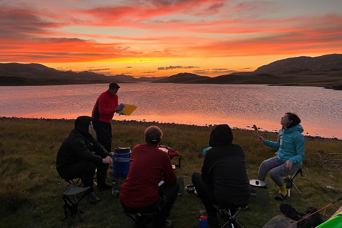 a group of adults doing a relaxing team challenge, sitting by a loch as the sun sets. beautiful orange skies behind