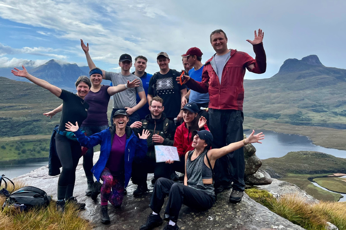a group of adults celebrating reaching the summit of a hill as part of a team challenge event. beautiful scenic landscape in background.