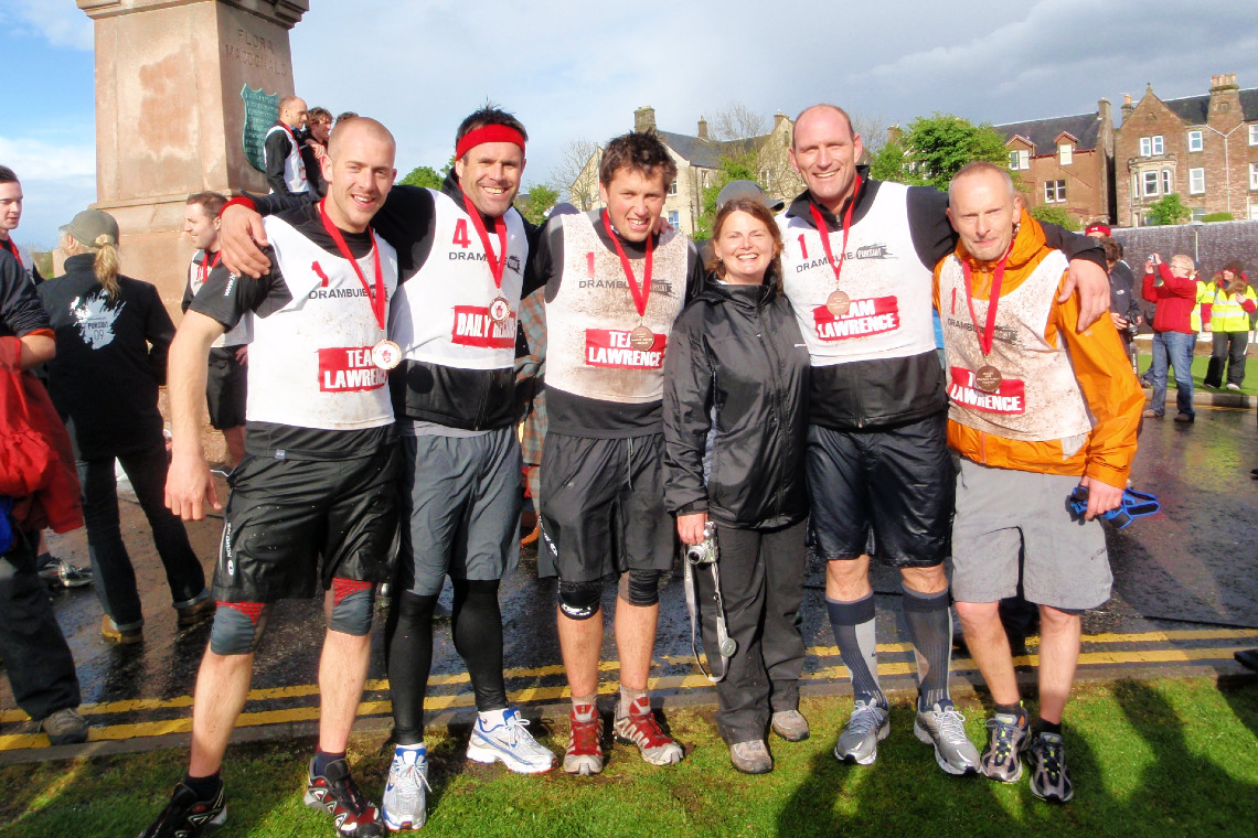 a group of adventure racers at the finish line celebrating with their medals. Event: Drambuie Pursuit (incl. Kenny Logan and Lawrence Dallaglio team captains)