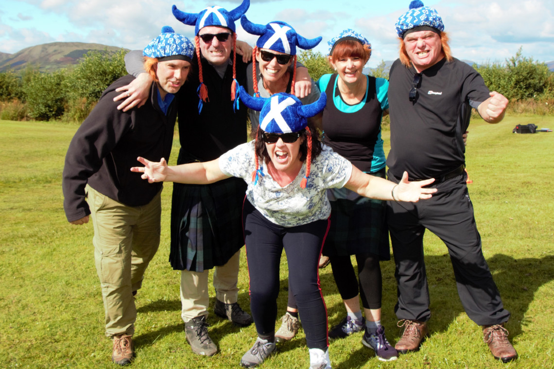 a group of adults, 3 of them in novelty scottish hats, cheering and smiling while posing for the camera, after a highland games event.