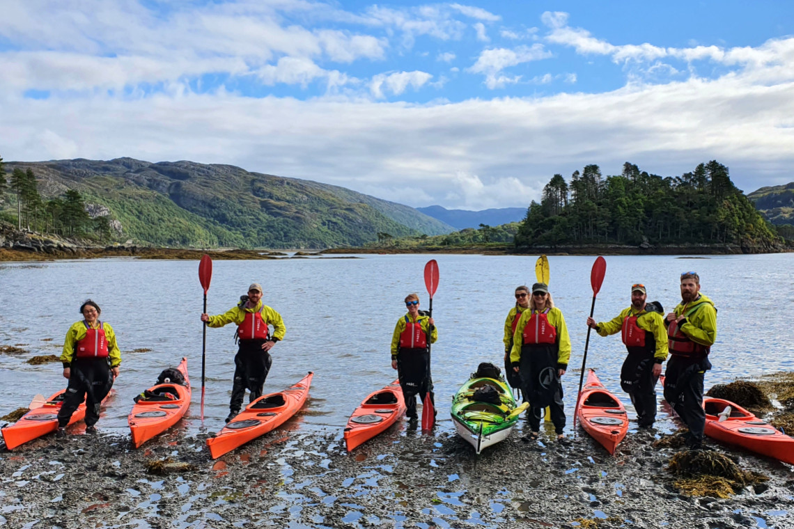 sea kayakers stand next to their kayaks, holding their paddles high, posing for the camer