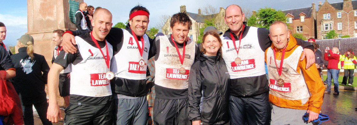a group of adventure racers at the finish line celebrating with their medals. Event: Drambuie Pursuit (incl. Kenny Logan and Lawrence Dallaglio team captains)