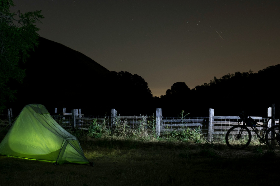 a tent iluminated by a light inside, against the back ground of a starry night sky. a bike is illuminted by another light to the right side of the photo.
