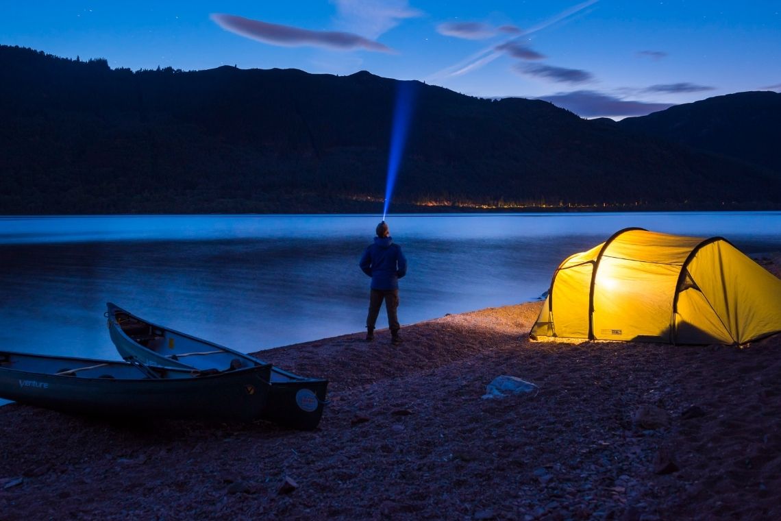 a person shining their head torch into the night sky, on the shore of a scottish loch. in the foreground is their tent illuminated from the inside by a light.