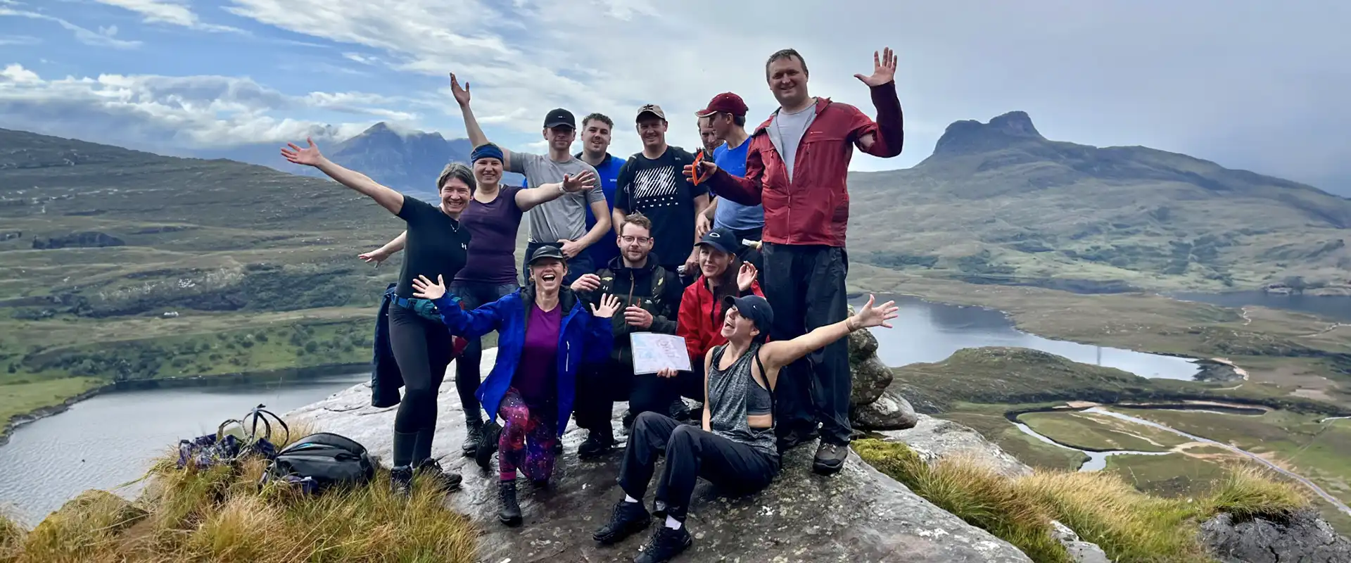 a group of smiling people posing for a camera on top of a hill with mountainous backdrop. Northwest highlands