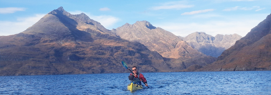 man sea kayaking on isle of skye coastline with cuillin mountains and blue skies in background