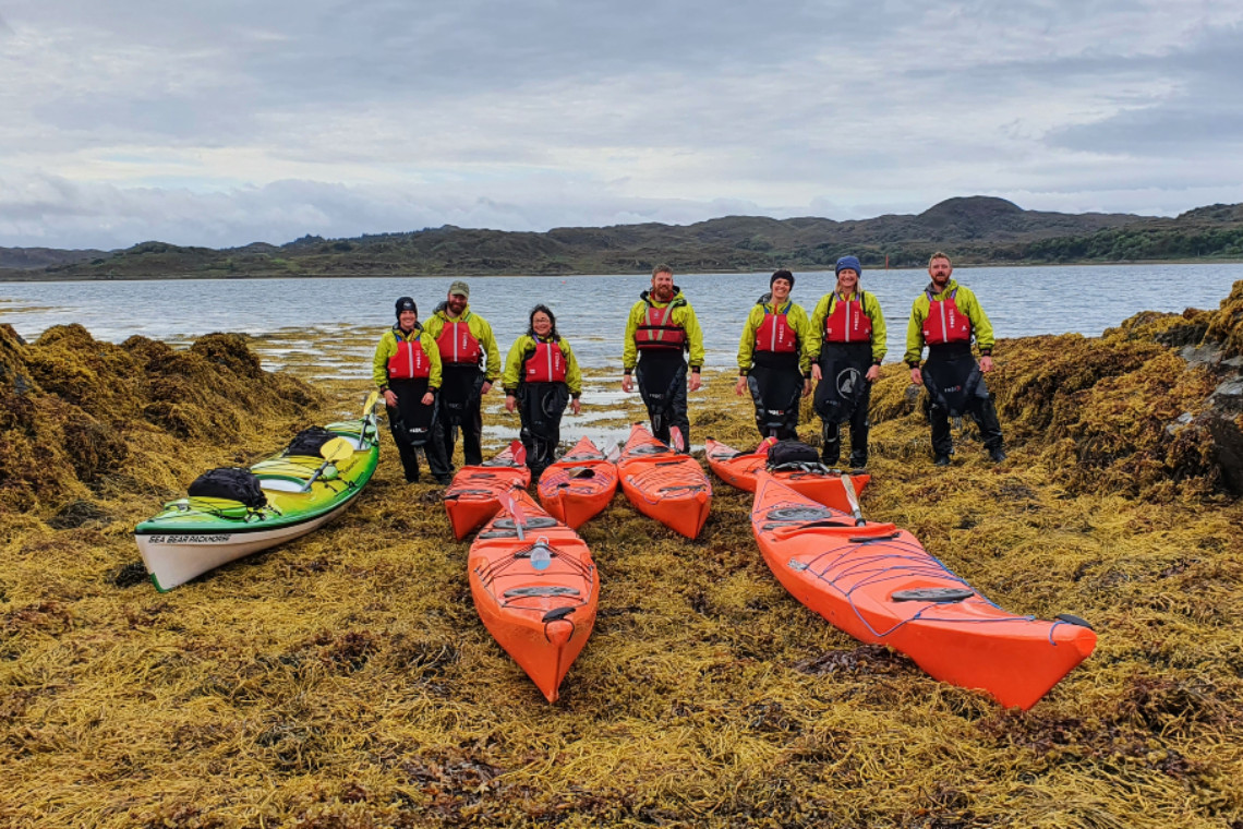 a group of people standing next to their sea kayaks on a beach covered in seaweed, the sea behind them