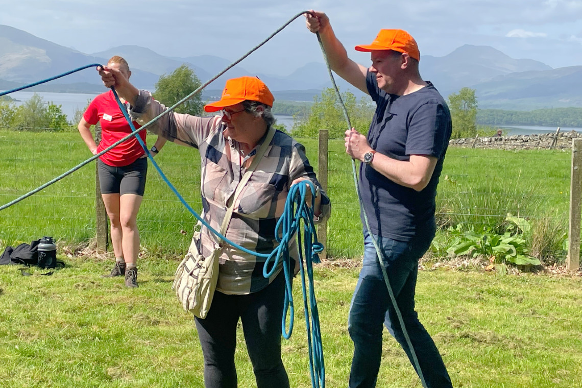 a lady and a man holding ropes above their heads while watching something else off photo. Part of a fun team challenge event. lovely view in the background to a loch and mountains.